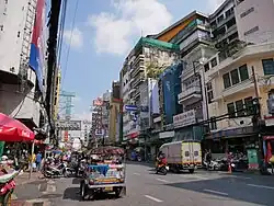 A street during daytime lined with many stalls and shops with a lot of signs bearing Thai and Chinese names