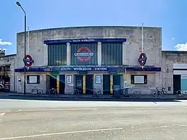 A grey building with a rectangular, dark blue sign reading "SOUTH WIMBLEDON STATION" in white letters all under a light blue sky