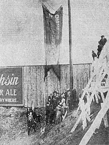 A black and white photograph of a flag hanging from a flag pole infront of a wooden outfield fence while several men and women stand below