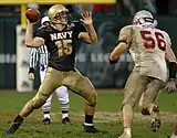 An American football player in a mud-covered blue and gold uniform prepares to throw a forward pass to another player out of frame. Another player in a white and red uniform leans forward to tackle him.