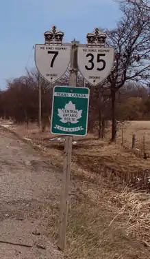 "An image of a signpost on a clear winter day. Behind the signpost are several leafless trees. To the left of the foot of the sign is the gravel shoulder of the road (not pictured) that it stands beside. The signpost is wooden, with three signs. Two are at the top, and one is centred below those. The top two are bullet-shaped signs with a king's crown on top. One is for Highway 7 and one for Highway 35. The sign below is green, with a white maple leaf in the centre. Above the leaf is a white banner with green text, reading "TRANS-CANADA HIGHWAY". The centre of the leaf reads in green "CENTRAL ONTARIO ROUTE". Finally, a white banner with a hung appearance is below the leaf. The green text within the banner reads "ONTARIO""