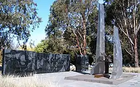 Monument consisting of two upright dark stones in front of a dark stone wall, with trees in the background