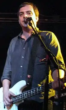 Adam Schlesinger is playing a guitar and singing live in front of a microphone.