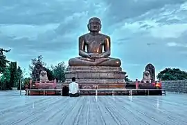 Large outdoor statue of Mahavira, with a seated worshipper for scale