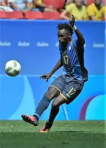 A man wearing navy blue shorts and a blue jersey kicking a football.