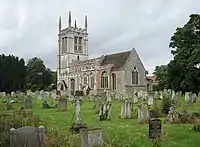 The tower of a stone church, seen from the west, at the top of which is a battlemented parapet with pinnacles.  In front of the church and to the left is a lychgate