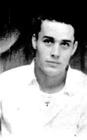 A black and white photograph of a man with dark hair leaning towards the camera slightly, wearing a light-colored shirt with the collar open, against a dark background