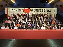 A table with seven steps of people with a banner ezker abertzalea in the background and the flags of Navarre on the left and the Basque Country on the right.