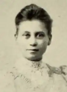 A young white woman with dark hair brushed back from her forehead, wearing a high-collared white lacy blouse or dress