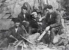 "Black and white photograph of a group of four IRA men in civilian clothing, one wearing a cap; they appear to be cooking in a rural location, as a large amount of wood can be seen in the foreground"