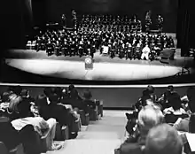 College graduates sitting on stage in a theater with spectators observing the ceremony in the foreground