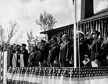 Men in suits and uniforms stand on a dais decorated with bunting and salute.