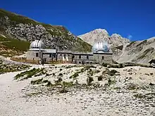 A rocky mountainous landscape has scrub brush scattered around the area. A stone building with two large silver domes is the observatory.