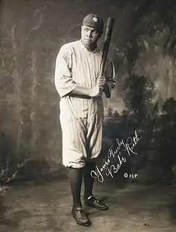 Full body shot of baseball player Babe Ruth, holding a bat and wearing a "NY" hat.