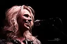 Close-up shot of a woman's face as she sings into a microphone. Her slightly, wavy, blonde hair falls to her shoulders.