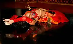 a woman in an elaborate red costume lies on her back with her legs folded under her, her arms above her head, and a sword balanced on her torso