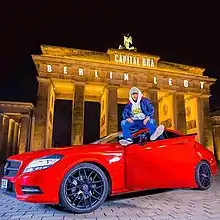 Capital Bra sitting on a red Mercedes-Benz in front of the Brandenburg Gate in Berlin.