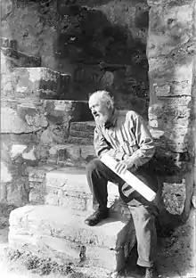 A black-and-white photograph of the architect Bernard Maybeck, dated 1919. In the photograph, Maybeck is gazing at the left side of the frame and resting on a step, slightly leaning to his right with his right foot drawn up on a lower step, the left foot on the ground, and the left hand holding a large rolled paper (possibly a blueprint, indicative of his work as an architect).