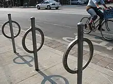 Unpainted steel rings, each welded to a steel post (running through it), set in concrete slabs. A cyclist is passing by from left to right on the cycle path behind.