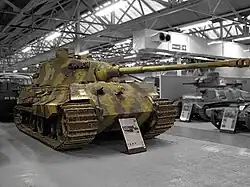 A large, turreted tank with dull yellow, green and brown wavy camouflage, on display inside Bovington museum. The tracks are wide, and the frontal armour is sloped. The long gun overhangs the bow by several meters.