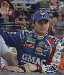 A man in his early-twenties wearing a blue-and-black baseball cap and a blue jacket with sponsors' logos; his right arm is elevated in a waving gesture.