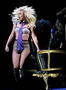 Image of a blond female performer. She has a headset around her hand and is wearing sparkly silver and black lingerie, fishnet stockings and knee-high black boots. She stands in front of a black and golden couch.