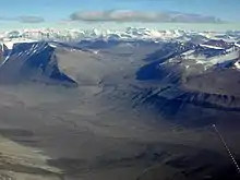  In the foreground is a landscape of dull brown mounds and undulations, behind which are snow-covered mountain peaks.