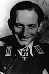 A black-and-white photograph of a smiling man wearing a military uniform and neck order in shape of an Iron Cross. His hair is combed back.