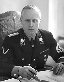 A blonde-haired male wearing a dark Nazi Party uniform with swastika armband seated at a desk