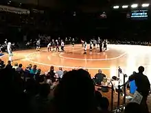 A flat wooden surface with roller skaters wearing helmets and knee pads standing on it. Some are wearing black and white striped shirts. People sit on the ground surrounding it and in bleachers.