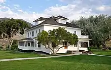 A white two-story house with front and side porches and a widow's walk rests on a property with green lawn, trees, and a picnic table.