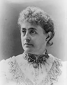 3/4 profile of the head and shoulders of a middle aged woman facing right with short hair in an elaborate white blouse with a highly beaded neckband and embellished with an embroidered yoke trimmed in decorative lace.