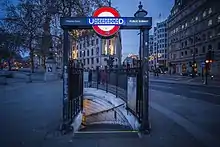 A narrow stair surrounded by decorative railings curves down from the pavement. The London Underground logo forms part of sign above. Large stone buildings line the street in the background