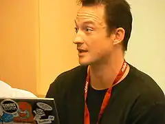 A Caucasian male sitting in front of a laptop. He has brown hair, a black shirt, and a red lanyard.