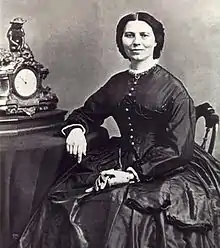 19th century photograph of a woman seated with her arm resting on a table. Her dark hair is neatly parted in the middle. She is smiling slightly