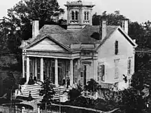 A white house with multiple chimneys and a tower.  People are on the front porch and someone is arriving at the home and about to embark up the staircase. The front porch has a roof over a set of four columns. Trees are visible on the lawn.