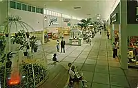 The interior of a shopping mall in the 1960s, with tiled floor. Visible are a fountain, several kiosk shops, planters with trees, and signage reading "Hanover Shoes", as well as several customers.