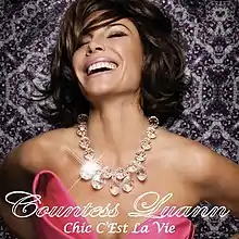 A headshot of Luann de Lesseps appears; she is shown wearing a pink dress and a large, transparent necklace.