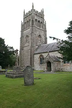 Stone building with arched porchway. Prominent square tower to the left hand end of building. In the foreground gravestones and crosses on grass.