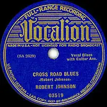 Photo of original 78 record label showing "Vocalion" in stylized lettering with "Cross Road Blues", "Vocal Blues with Guitar Acc[ompaniment]", and "Robert Johnson" as identifiers along with catalog numbers, "Made in USA", etc.