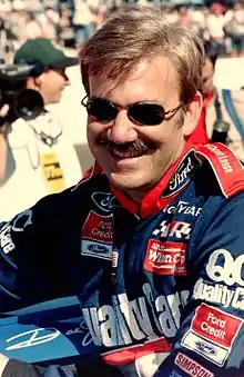 A mustached man in his early forties wearing sunglasses and blue and red racing overalls