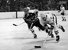 Two hockey players in full pads and helmets on the ice, both in motion, with two others further behind them.