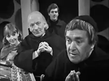 The Monk (right) looks eagerly to the left of the camera. In the background is the Doctor (dressed in monk clothing), Vicki, and Steven. They are standing in the Monk's TARDIS at the main console; the roundel walls can be seen in the far background, and the doors are open.