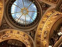 Photo showing the ceiling in the domed hall. The ceiling is intricately decorated with the glass dome and gold details on the arches.