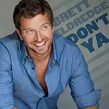 A smiling man wearing a sleeved-up blue shirt with the top button opened.