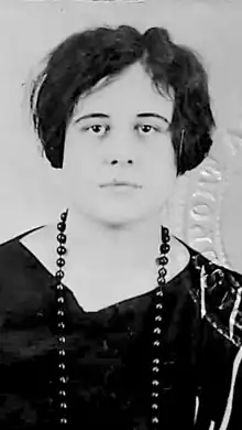 A young white woman with tousled dark cropped hair, wearing a v-neck dark top and dark beads; the edge of an embossed seal is visible in the image