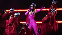 Dua Lipa performing in a pink catsuit surrounded by dancers in red outfits holding disco ball-styled hearts