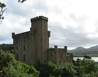 A grey castle with tall square towers stands amongst trees in full leaf