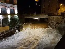 Night shot showing the Durolle in flood, with a very large flow of brownish water at a small waterfall in the middle of town, with a factory on the bank to the left of the shot.