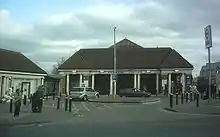 A white bluilding with white columns upholding a brown-shingled roof with people walking and standing in front all under a blue sky